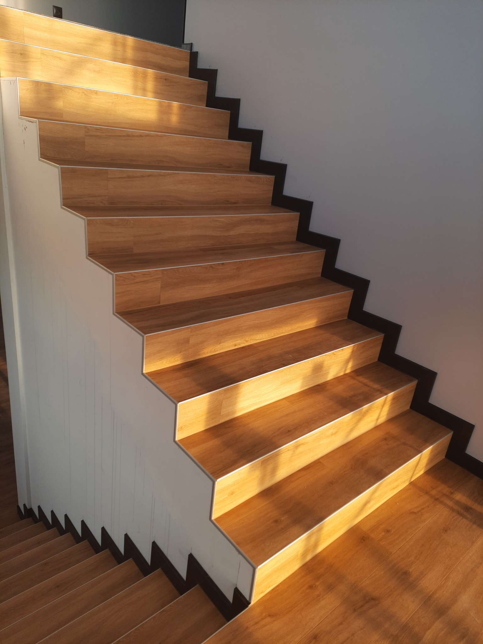 Schody wykończone panelami drewnopodobnymi z aluminiowymi listwami krawędziowymi, widok z góry w dół, oświetlone ciepłym światłem słonecznym.