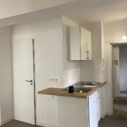 Mała, biała kuchnia z drewnianym blatem, zintegrowana z małą lodówką i płytą indukcyjną, widok na białe drzwi i szary podłogę.