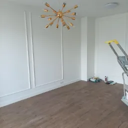 Pomalowane na biało wnętrze z drewnopodobną podłogą, sztukaterią ścienną i złotą lampą w stylu retro; drabina malarska i akcesoria malarskie w rogu.