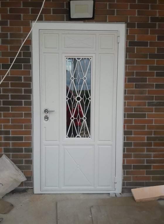 Białe drzwi wejściowe z dekoracyjnym przeszkleniem, zamontowane w elewacji z cegły, oświetlone zewnętrzną lampą.