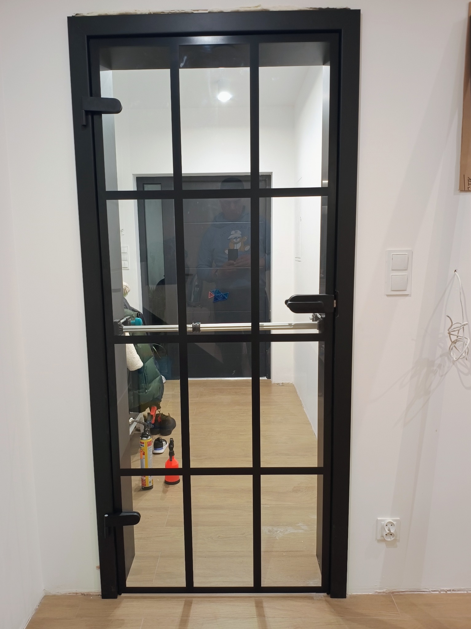 Czarna, szklana drzwi wewnętrzne z podziałami, widok na korytarz z narzędziami i odzieżą, białe ściany, drewniana podłoga.