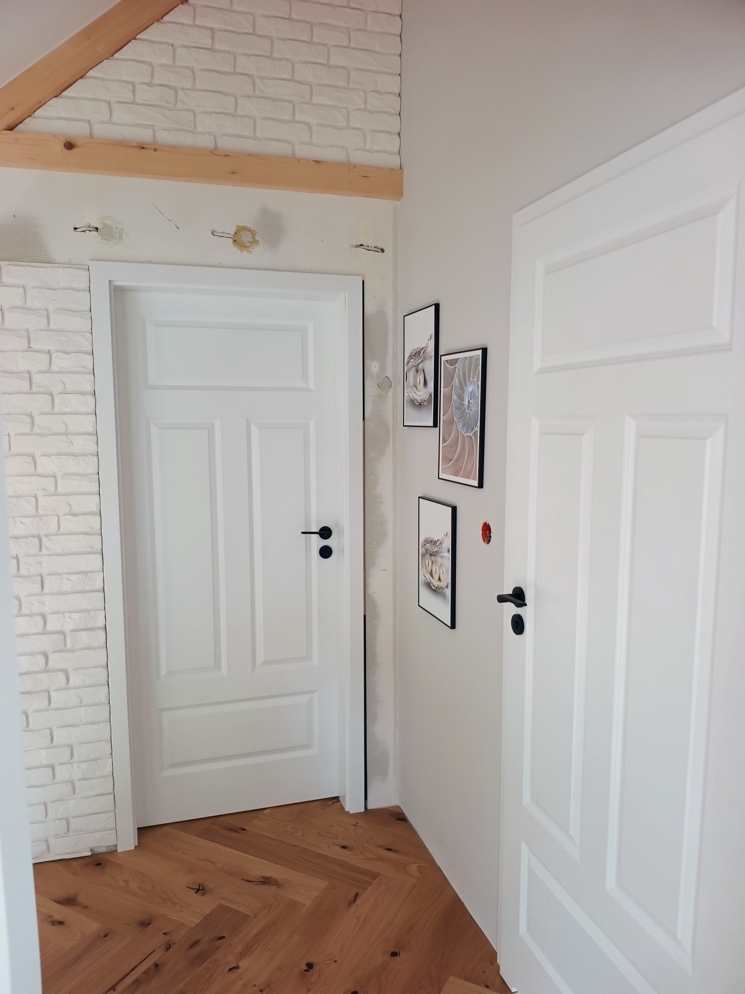 Białe drzwi wewnętrzne z czarną klamką w nowym domu, z drewnianą podłogą w jodełkę i dekoracjami ściennymi, widoczne ślady po niedokończonych pracach wykończeniowych.