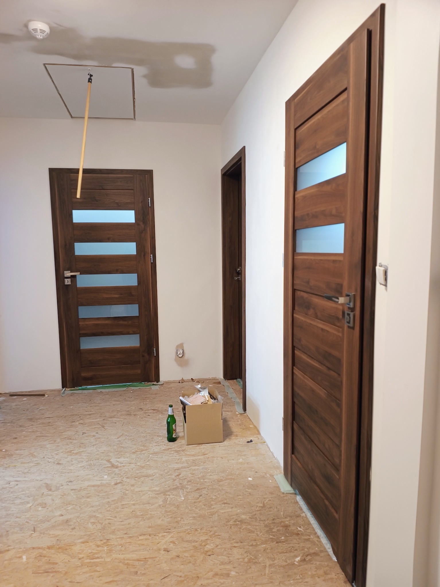 Dwie drewniane drzwi wewnętrzne z przeszkleniami, jedna zamontowana, druga w głębi, widoczna butelka i karton na podłodze z płyt OSB, sufit z otworem rewizyjnym.