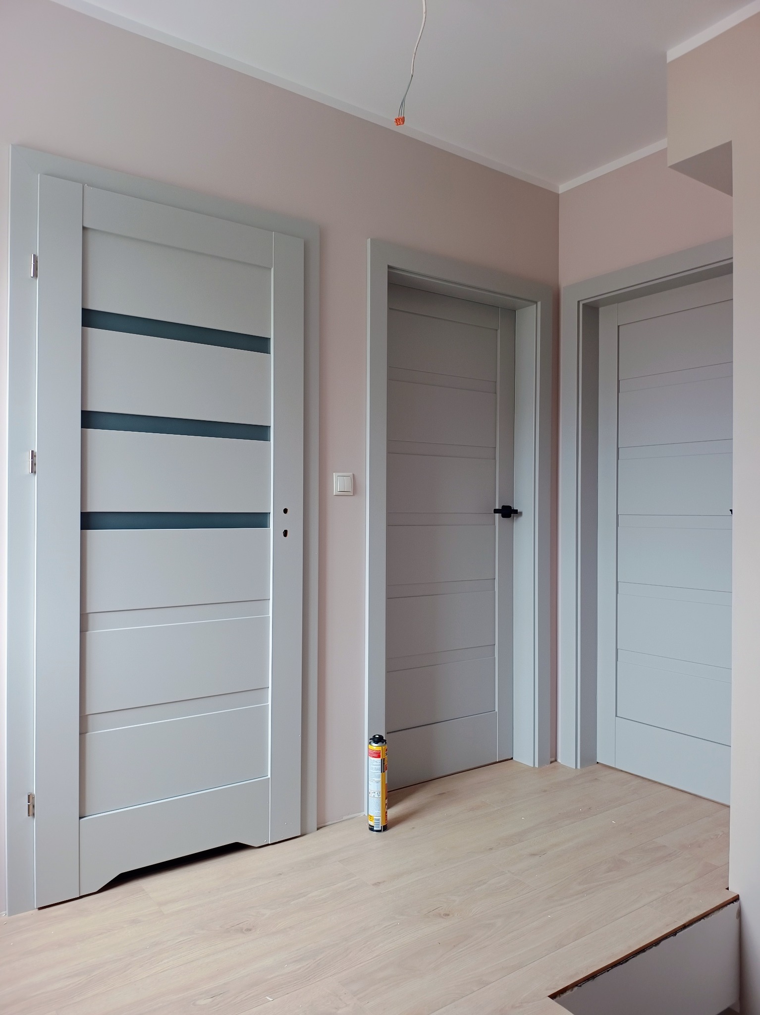 Wnętrze z trzema szarymi drzwiami wewnętrznymi o różnych wzorach, z widoczną pianką montażową przy jednej z nich, na podłodze panele.