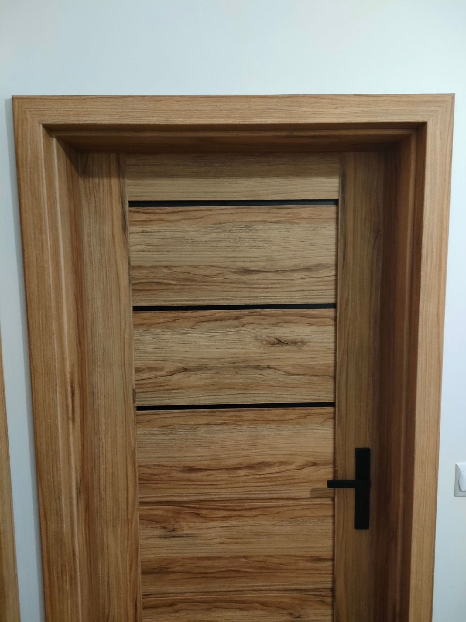 Drewniane drzwi wewnętrzne z trzema poziomymi, czarnymi intarsjami i czarną klamką na tle białej ściany.