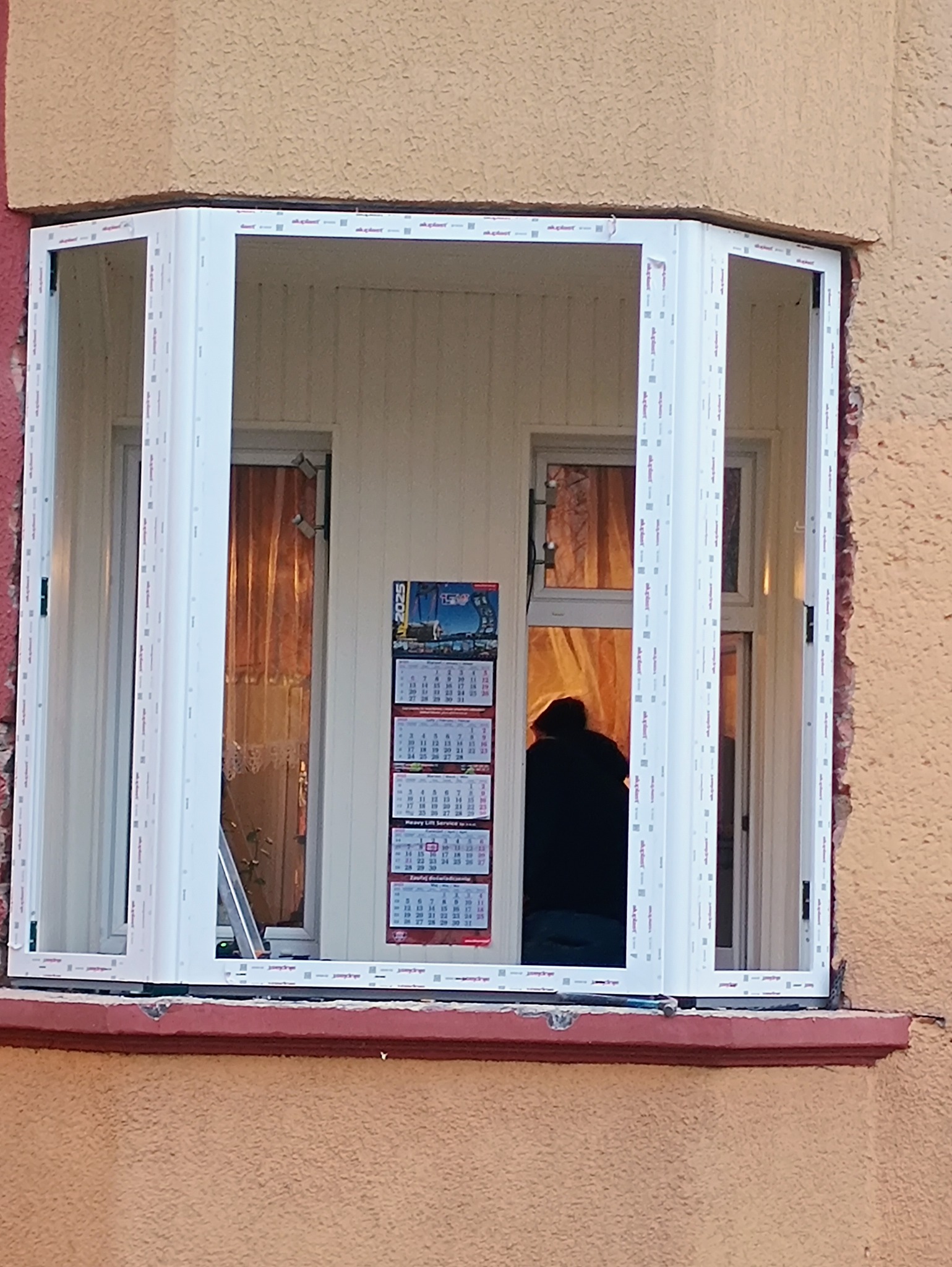 Wstawiony szkielet okna PCV w wykusz, widoczne fragmenty muru i wnętrze z kalendarzem na ścianie.