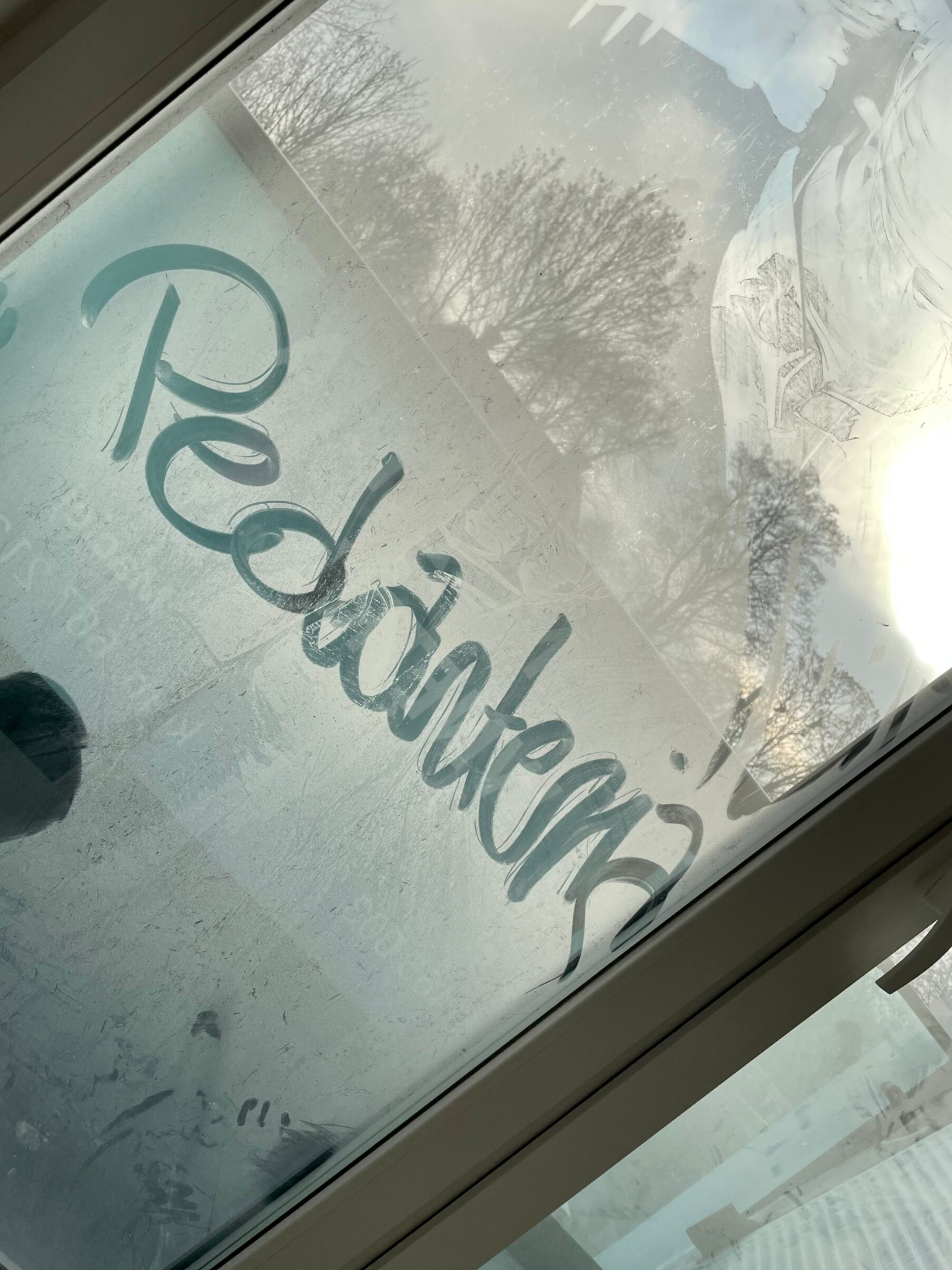 Brudne okno dachowe z napisem 'Redakcja' wykonanym palcem, widoczne zacieki i odbicie drzew na zewnątrz.