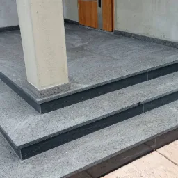 Zewnętrzne schody wejściowe z szarego kamienia z ciemniejszymi podstopnicami, prowadzące do drzwi wejściowych i kolumny podtrzymującej zadaszenie.