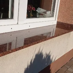 Polerowany, brązowo-czerwony parapet z kamienia naturalnego, zamontowany pod oknem z białymi ramami, odbijający światło i elementy wnętrza.