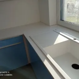 Biały blat kuchenny z wbudowanym zlewozmywakiem i otworami na baterię, zamontowany przy oknie, z niebieskimi szafkami poniżej.