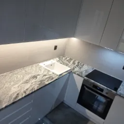 Szary blat kuchenny z kamienia o wyraźnym wzorze, wkomponowany w nowoczesną, białą zabudowę kuchenną z wbudowanym zlewem i płytą grzewczą.