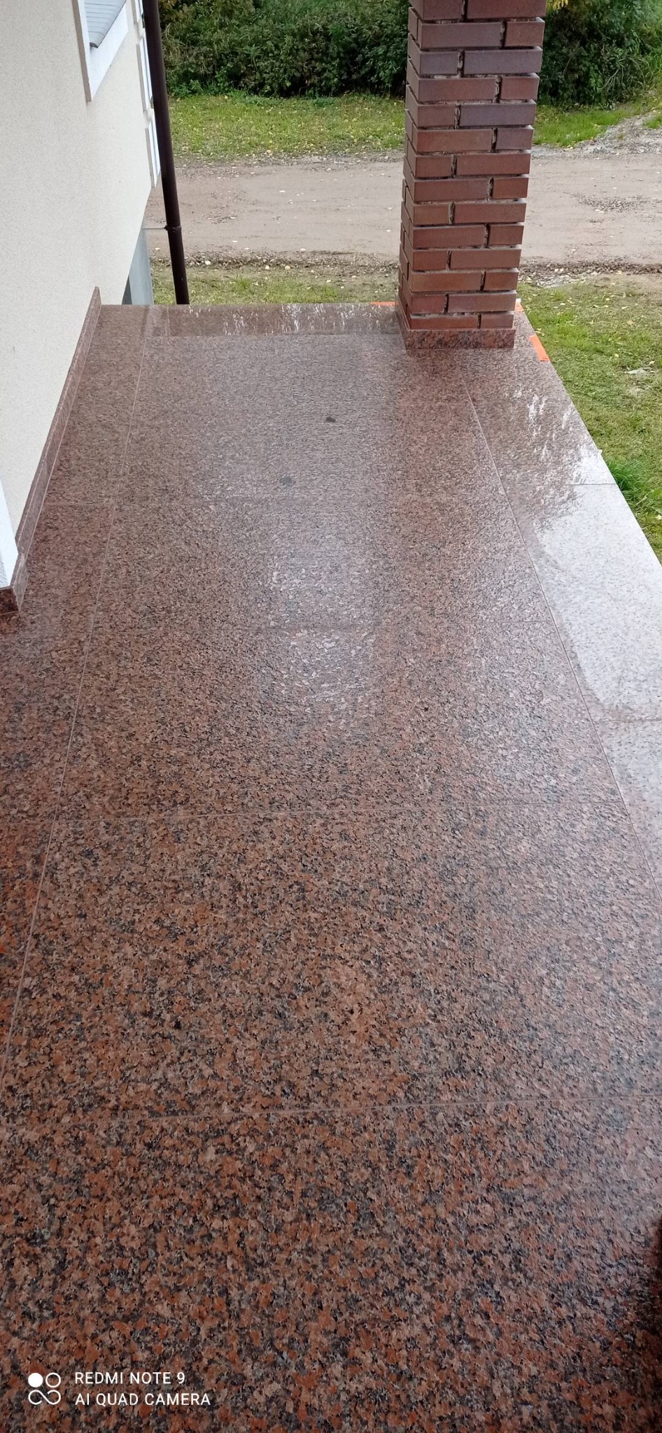 Wyłożony czerwonobrązowym, nakrapianym kamieniem taras z ceglaną kolumną podtrzymującą zadaszenie, widok z góry w pochmurny dzień.