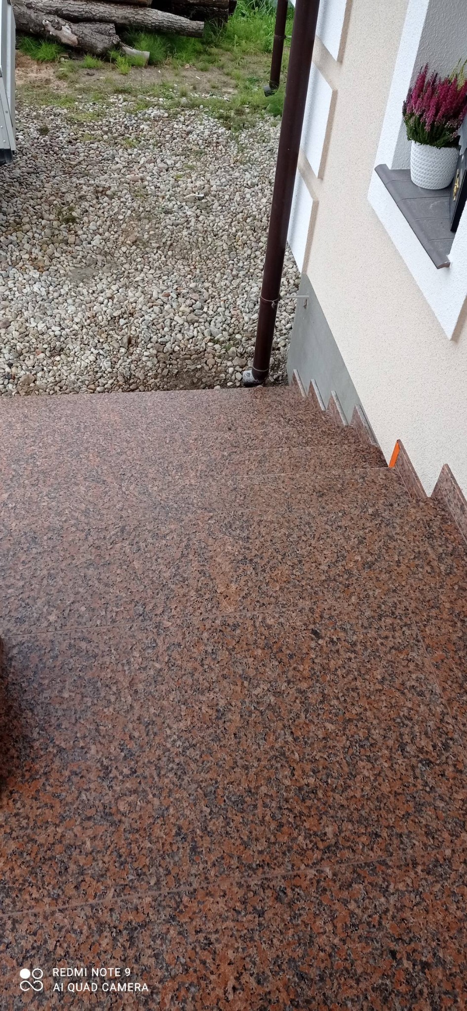 Zewnętrzne schody wejściowe wyłożone płytami z czerwono-brązowego granitu, widok z góry na tle żwirowego podłoża i fragmentu elewacji budynku z parapetem i kwiatami.