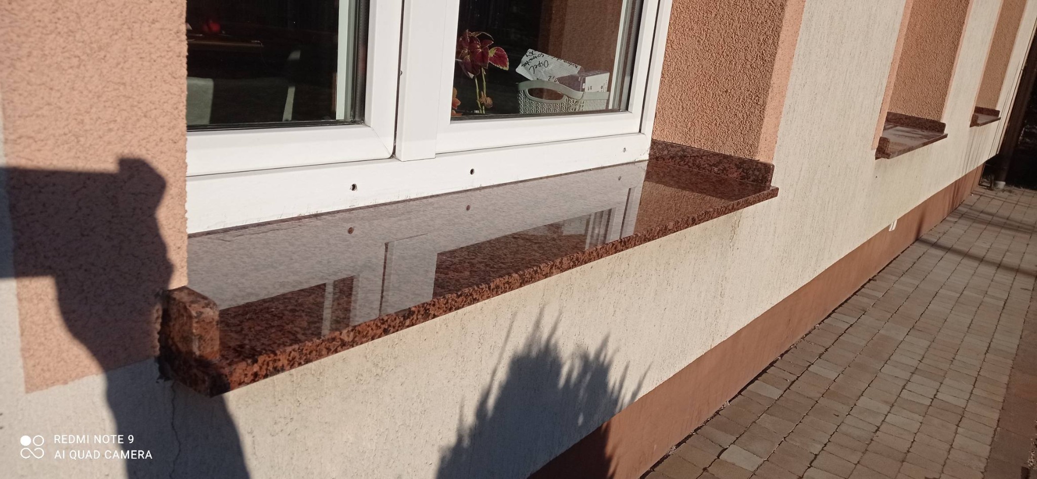 Polerowany, brązowo-czerwony parapet z kamienia naturalnego, zamontowany pod oknem z białymi ramami, odbijający światło i elementy wnętrza.