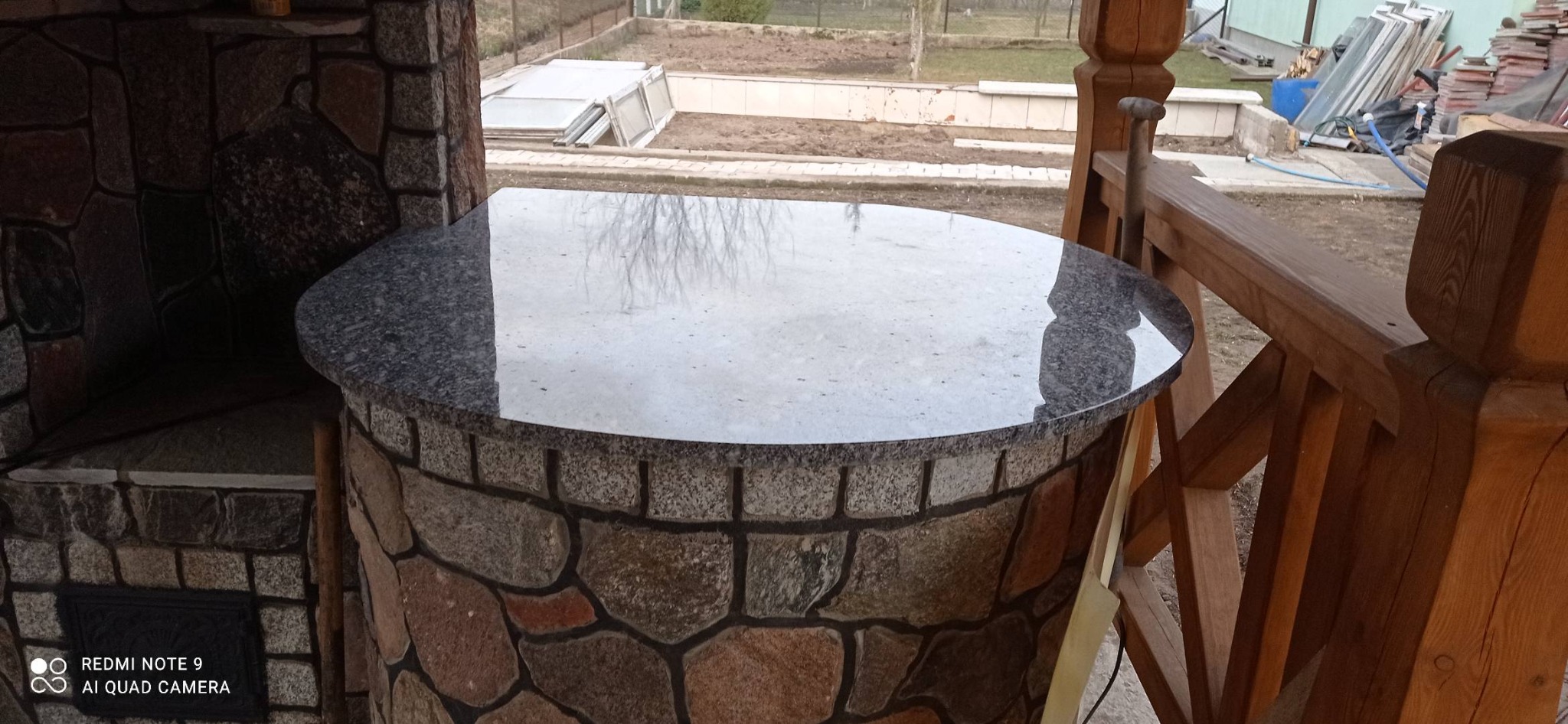 Okrągły blat z szarego kamienia na murowanym grillu zewnętrznym w altanie, odbicie drzewa na powierzchni.