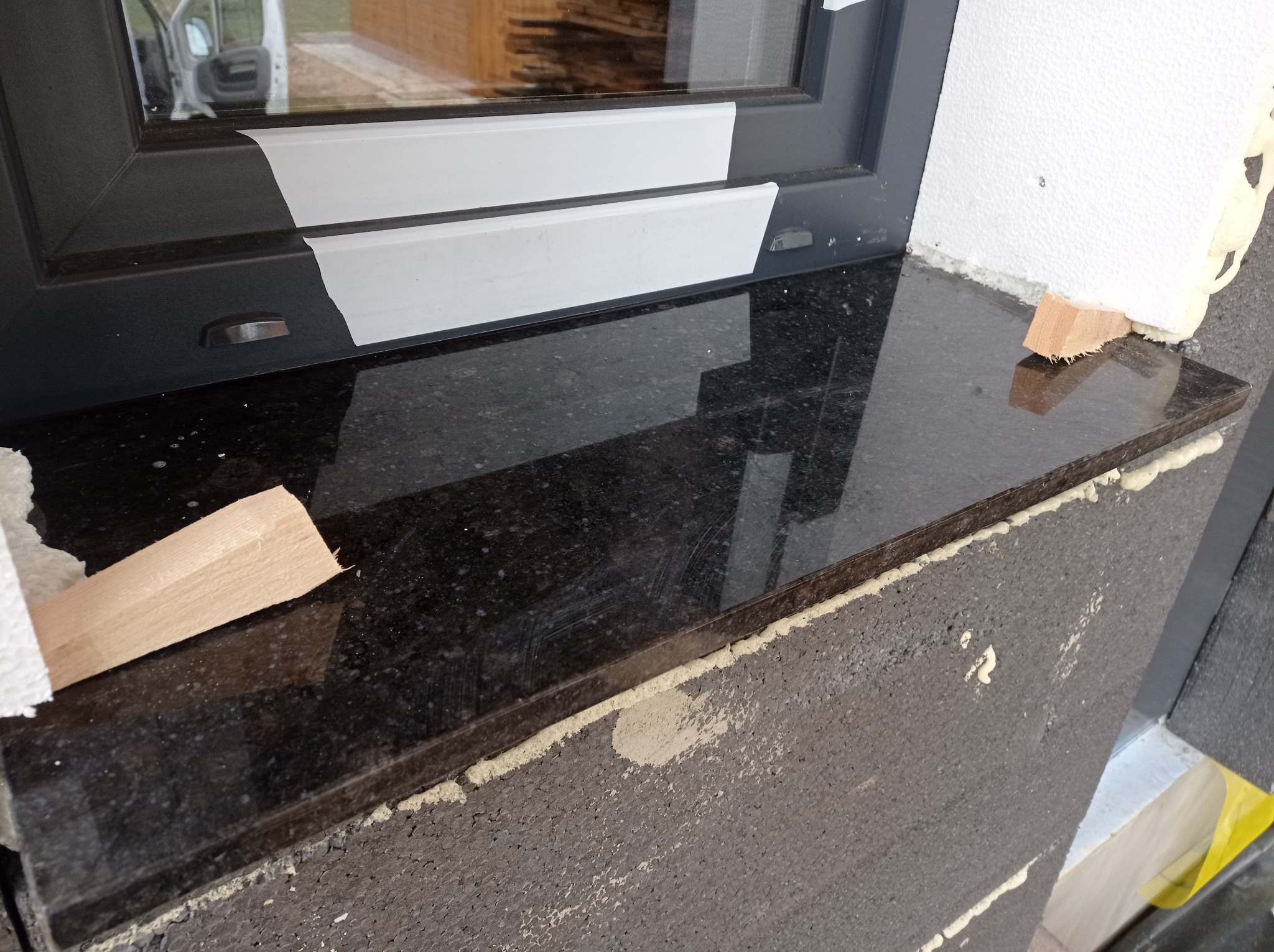 Montaż czarnego parapetu zewnętrznego z kamienia, z widocznymi klinami stabilizującymi i pianką montażową, osadzony w ociepleniu budynku.