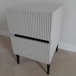 Biała szafka nocna z frezowanymi frontami szuflad i czarnymi nóżkami, ustawiona na parkiecie w jodełkę.