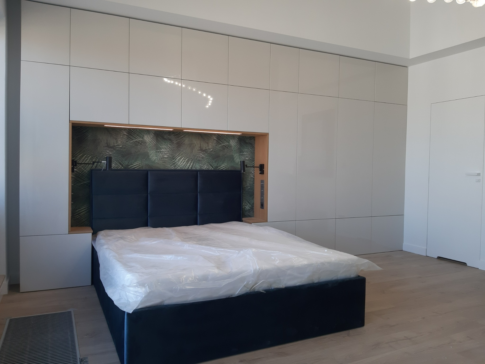 Białe szafy na całej ścianie z wnęką nad wezgłowiem łóżka, podświetlenie LED, wzorzysta tapeta we wnęce, łóżko z ciemnym obiciem przykryte folią ochronną.