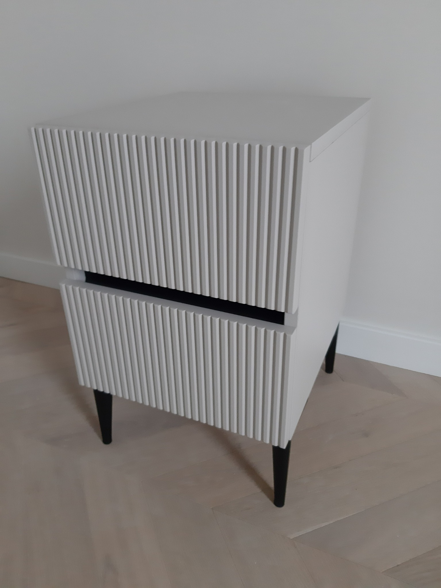 Biała szafka nocna z frezowanymi frontami szuflad i czarnymi nóżkami, ustawiona na parkiecie w jodełkę.