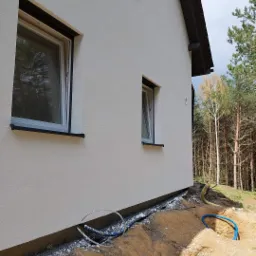 Świeżo otynkowana ściana domu jednorodzinnego z czarnymi obramowaniami okien, widoczne prace ziemne wokół fundamentów i wystające rury instalacyjne w wykopie.
