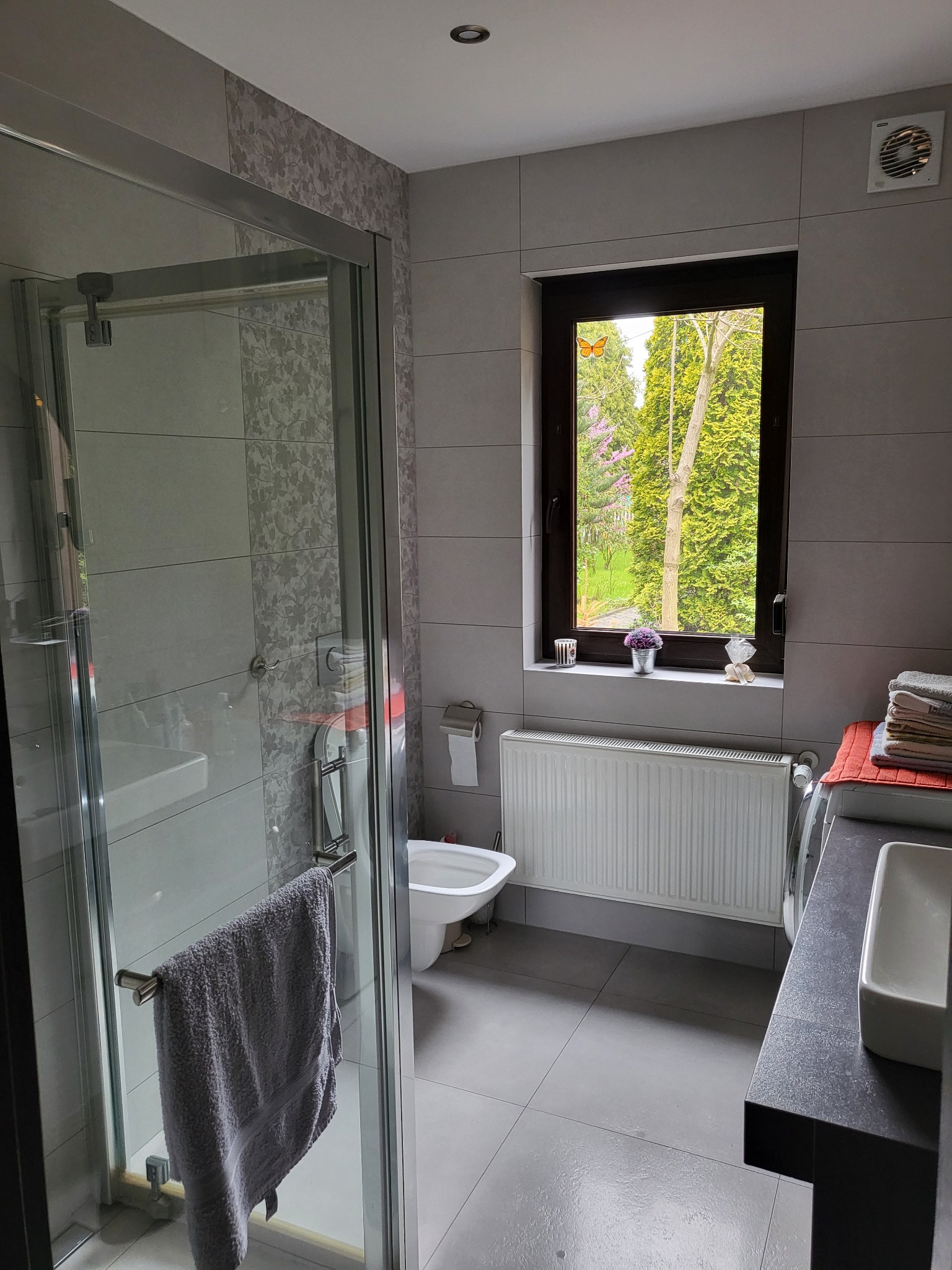 Jasna łazienka z szarą glazurą, kabiną prysznicową z szarą zawieszoną szmatką, oknem z widokiem na zieleń i dekoracjami, grzejnikiem i umywalką na blacie.