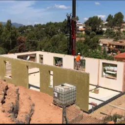 Montaż ściany modułowej domu z oknami przy użyciu dźwigu na tle zieleni i zabudowań mieszkalnych; pracownik na drabinie w pomarańczowej koszulce.