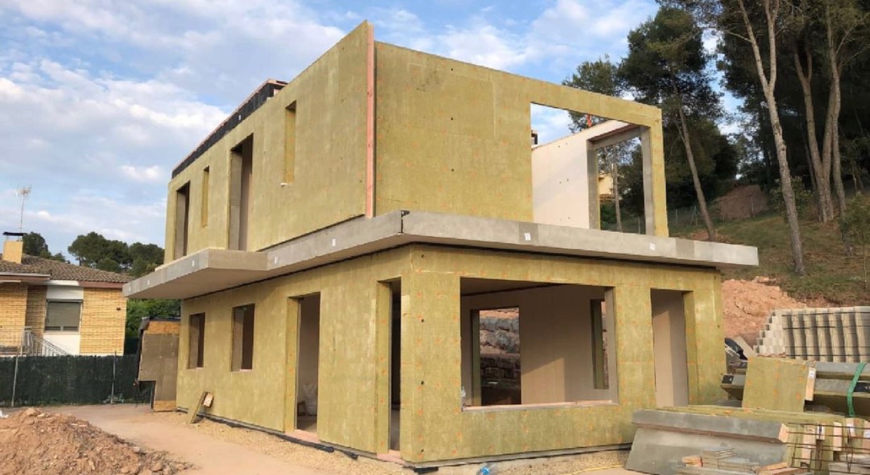 Dwukondygnacyjny budynek w trakcie budowy, z widocznymi panelami izolacyjnymi i betonowymi elementami konstrukcyjnymi, na tle drzew i nieba.