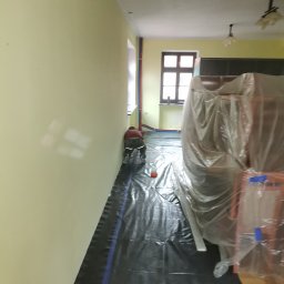 Magda Rzeszutek - Przygotowanie do remontu pokoju: pracownik zabezpiecza podłogę czarną folią malarską, meble owinięte folią ochronną, widoczne rury grzewcze przy ścianie.