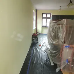Przygotowanie do remontu pokoju: pracownik zabezpiecza podłogę czarną folią malarską, meble owinięte folią ochronną, widoczne rury grzewcze przy ścianie.
