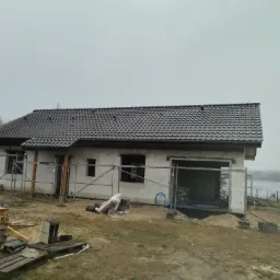 Surowy stan domu z białego pustaka z ciemną dachówką, otoczonego rusztowaniami i paletami, z widokiem na jezioro w tle.