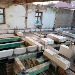 Magda Rzeszutek - Wnętrze budynku w trakcie rozbudowy: murowanie ścian z bloczków betonowych na drewnianej konstrukcji stropu, widoczne stare mury z oknami i przygotowane materiały budowlane.