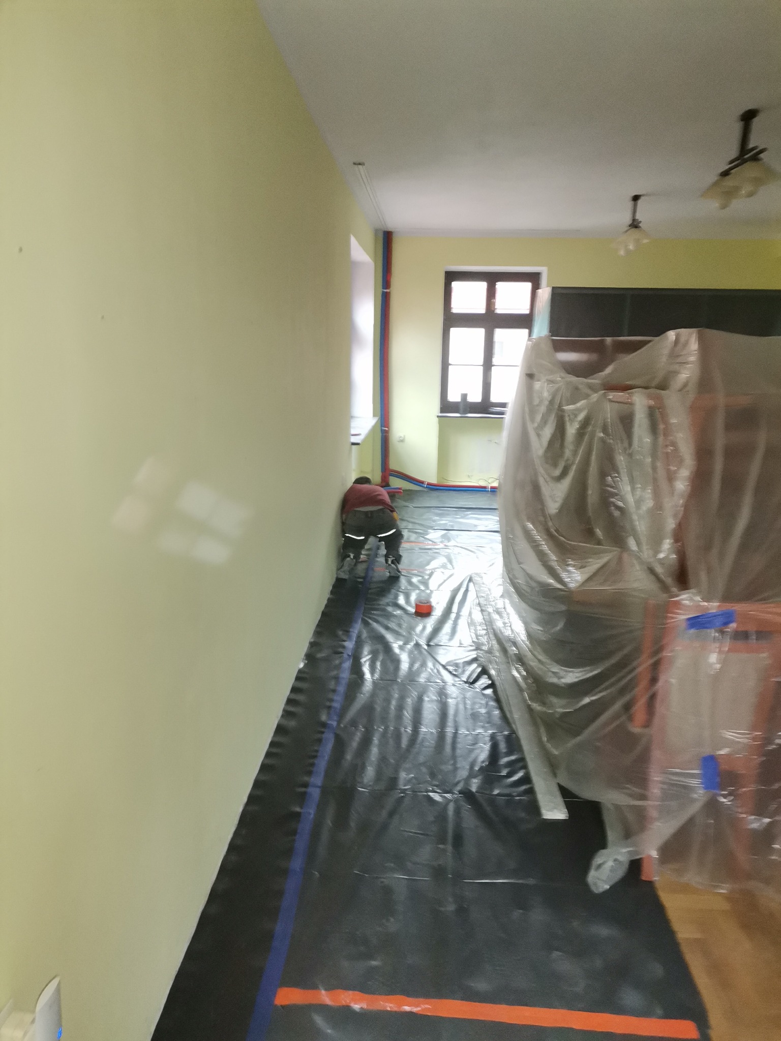 Przygotowanie do remontu pokoju: pracownik zabezpiecza podłogę czarną folią malarską, meble owinięte folią ochronną, widoczne rury grzewcze przy ścianie.