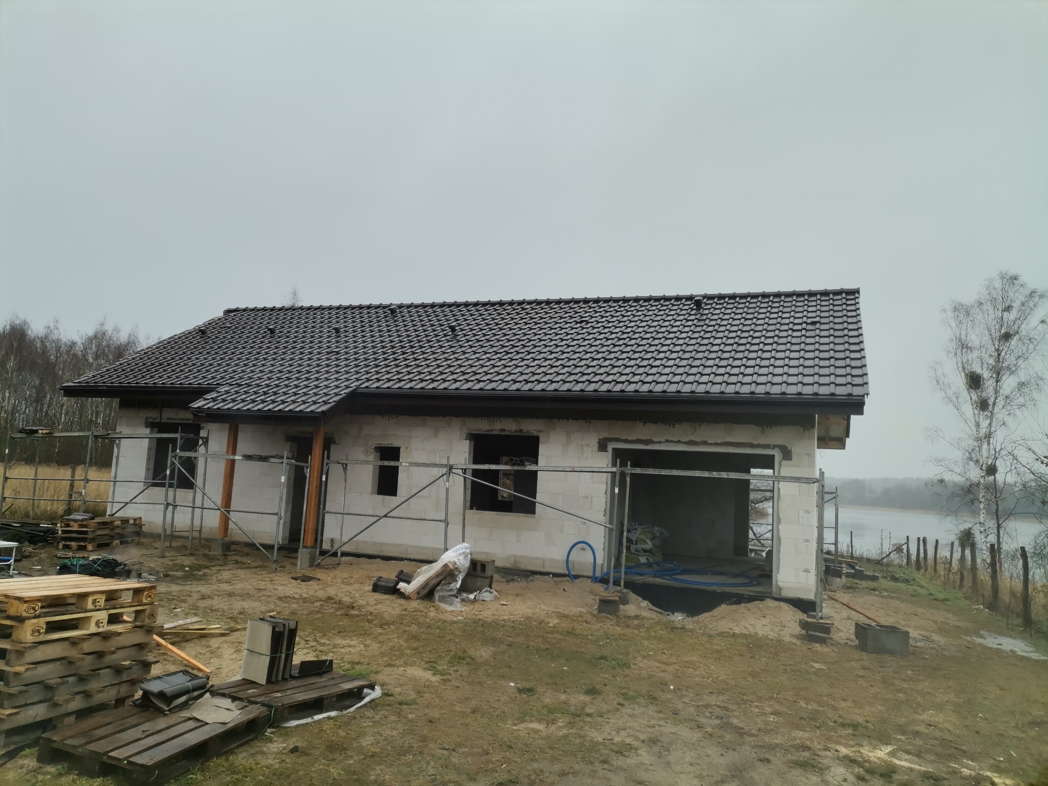 Surowy stan domu z białego pustaka z ciemną dachówką, otoczonego rusztowaniami i paletami, z widokiem na jezioro w tle.