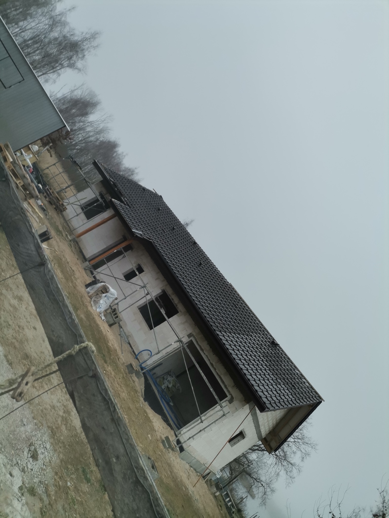 Surowy stan domu z białego pustaka z ciemną dachówką, widoczne rusztowania i przygotowany teren wokół budynku w pochmurny dzień.