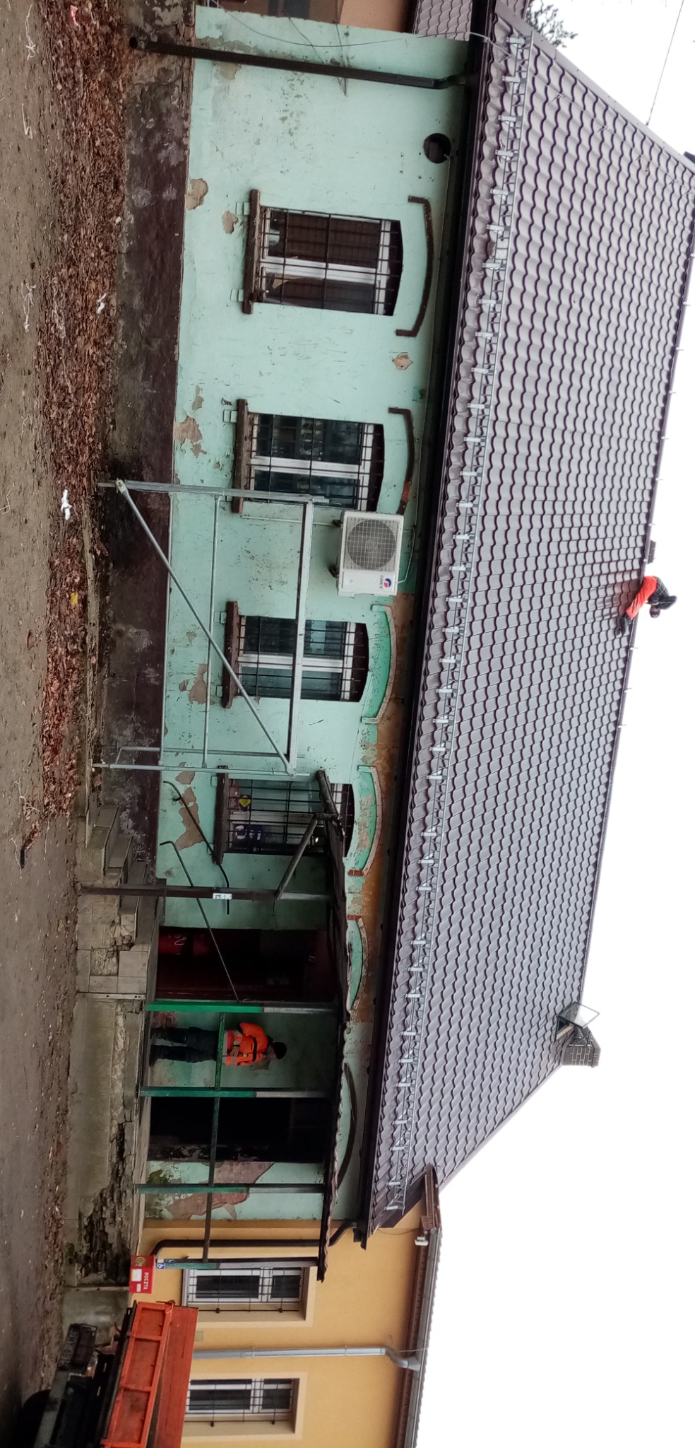 Pracownik w pomarańczowej kamizelce na dachu dwupiętrowego domu z blachodachówką, widoczne rusztowanie i odpadający tynk na elewacji budynku.