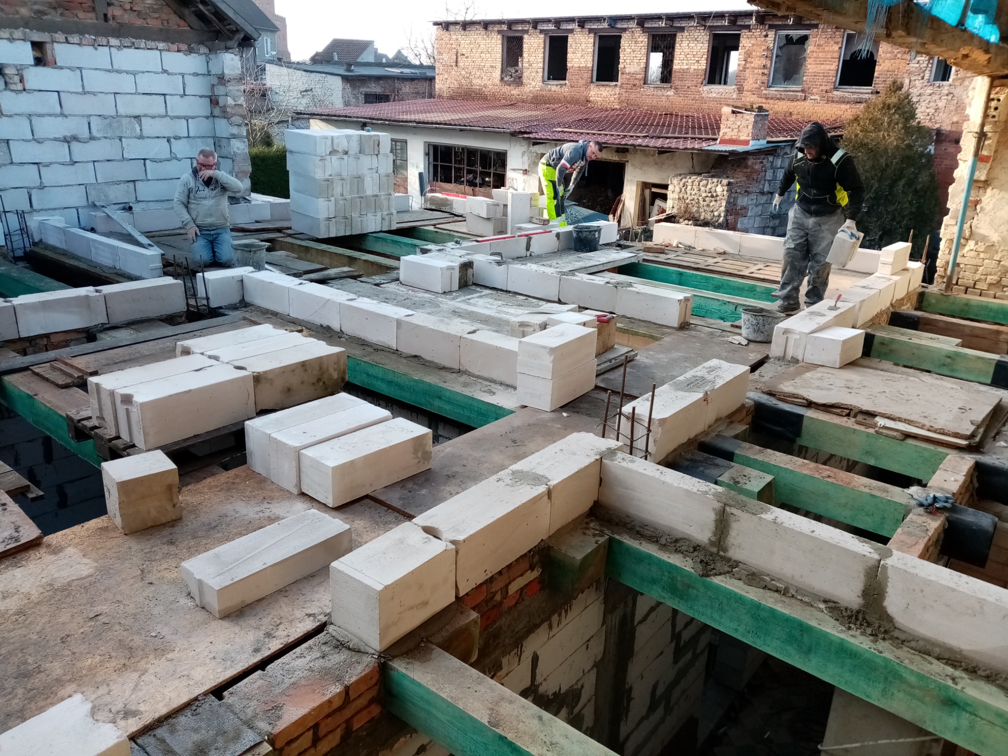 Trzech pracowników budowlanych na budowie domu z betonu komórkowego, widok z góry na wznoszone ściany i przygotowane bloczki. W tle zabudowa mieszkalna.