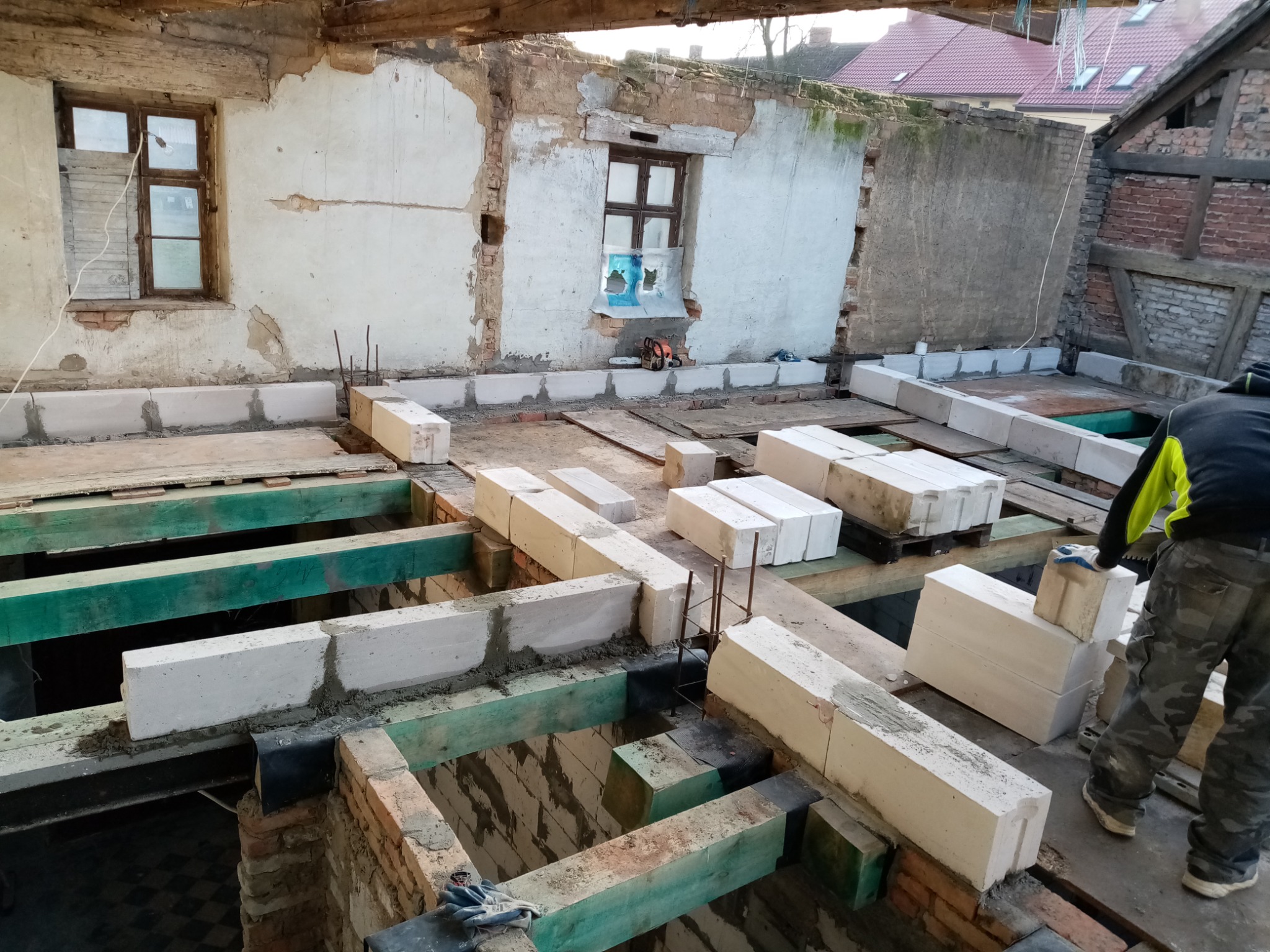 Wnętrze budynku w trakcie rozbudowy: murowanie ścian z bloczków betonowych na drewnianej konstrukcji stropu, widoczne stare mury z oknami i przygotowane materiały budowlane.