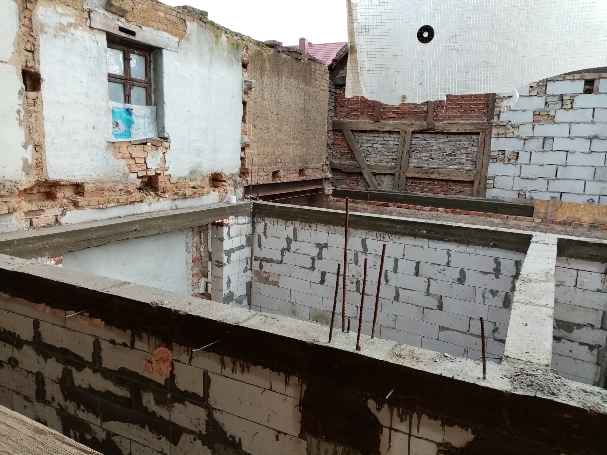 Budowa domu: widok na fundamenty i ściany w trakcie murowania z betonu komórkowego. Widoczne wystające pręty zbrojeniowe i fragmenty starej konstrukcji.