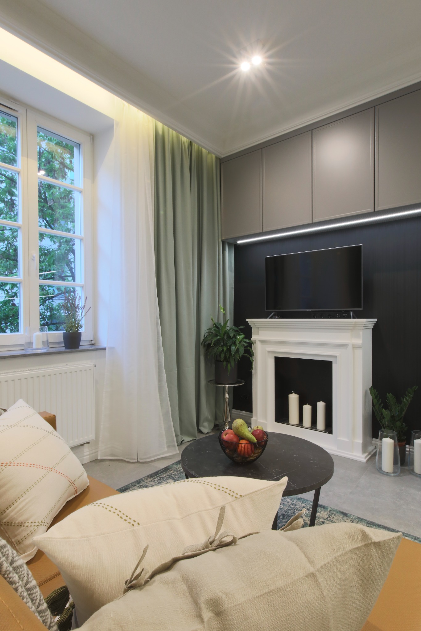 Salon z dekoracyjnym kominkiem, telewizorem, szarymi szafkami pod sufitem z oświetleniem LED, okrągłym stolikiem kawowym z owocami, beżową sofą z poduszkami i zasłonami w odcieniach bieli i zieleni.
