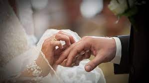 Zbliżenie dłoni pary młodej podczas ceremonii ślubnej; panna młoda zakłada obrączkę na palec pana młodego, widoczne detale sukni z perłami i złota obrączka z delikatnym wzorem.