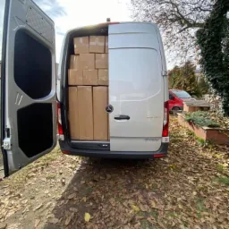 Srebrny van Mercedes Sprinter z otwartymi tylnymi drzwiami, wypełniony kartonowymi pudłami, widok z tyłu na tle jesiennego krajobrazu.