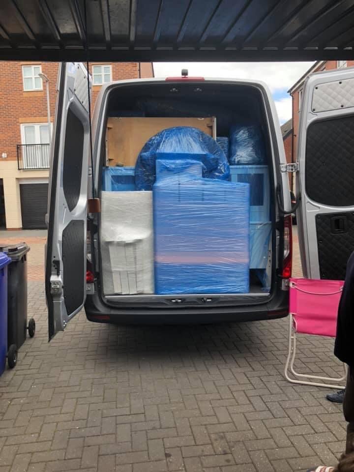 Wnętrze busa załadowane meblami owiniętymi folią stretch, przygotowanymi do transportu, widok z zewnątrz na tle brukowanej nawierzchni i budynków mieszkalnych.