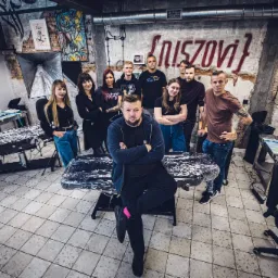 Grupowe zdjęcie zespołu w studiu tatuażu z widocznym sprzętem, lampami oświetleniowymi, stołami pokrytymi folią ochronną i dekoracjami na ścianach z surowej cegły.