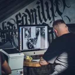 Grafik pracuje nad projektem na komputerze iMac, widoczne oprogramowanie graficzne z edytowaną grafiką w stylu tatuażu, w tle graffiti na ścianie i drukarka.