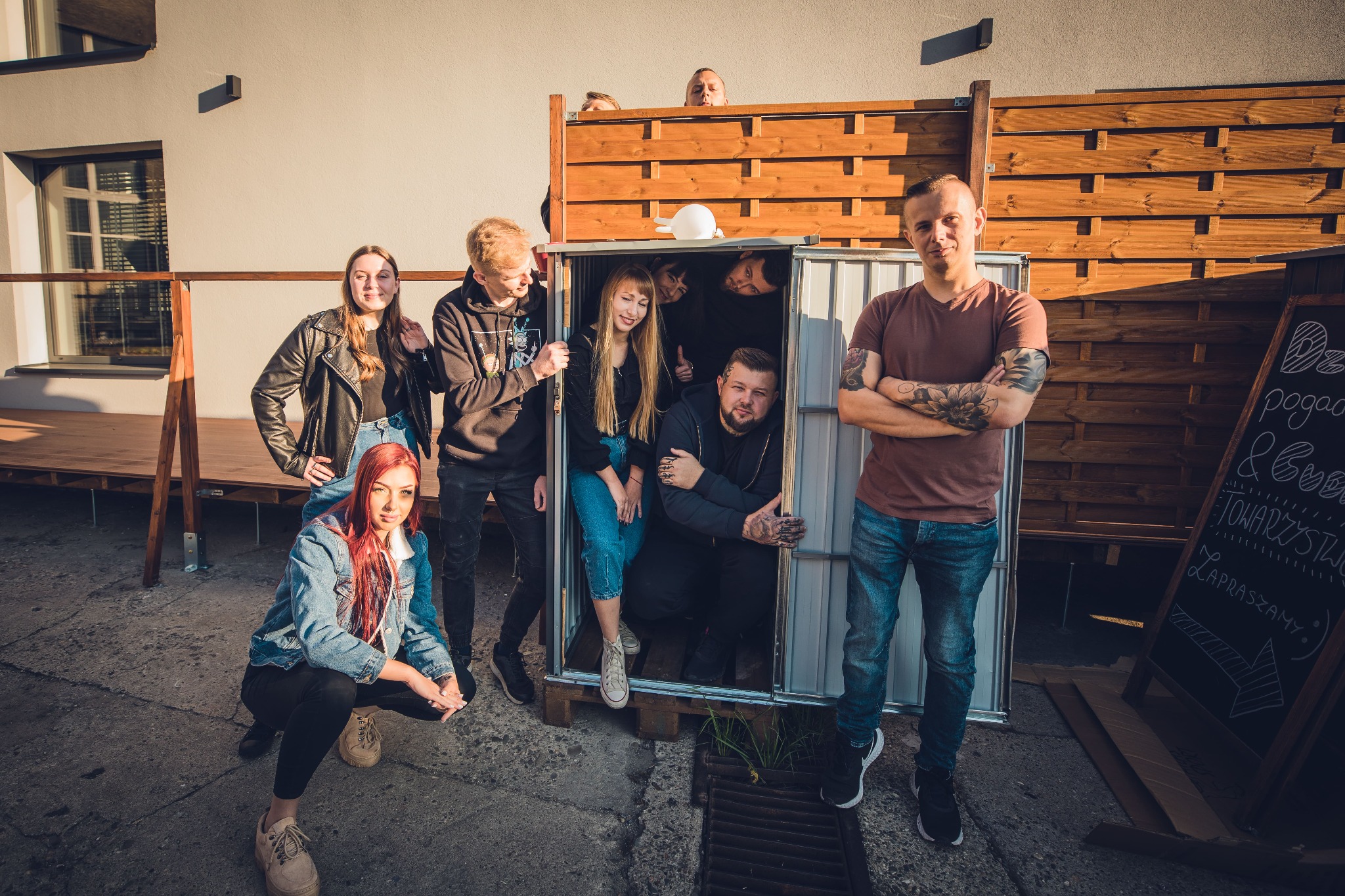 Grupa osób pozuje do zdjęcia, niektórzy wewnątrz metalowej konstrukcji przypominającej budkę, na zewnątrz budynku z tablicą informacyjną.