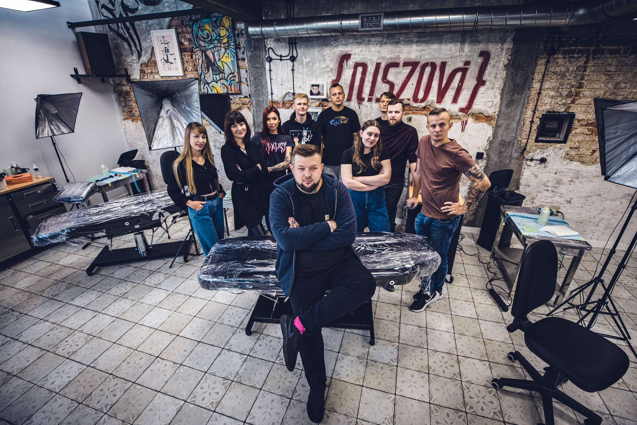 Grupowe zdjęcie zespołu w studiu tatuażu z widocznym sprzętem, lampami oświetleniowymi, stołami pokrytymi folią ochronną i dekoracjami na ścianach z surowej cegły.