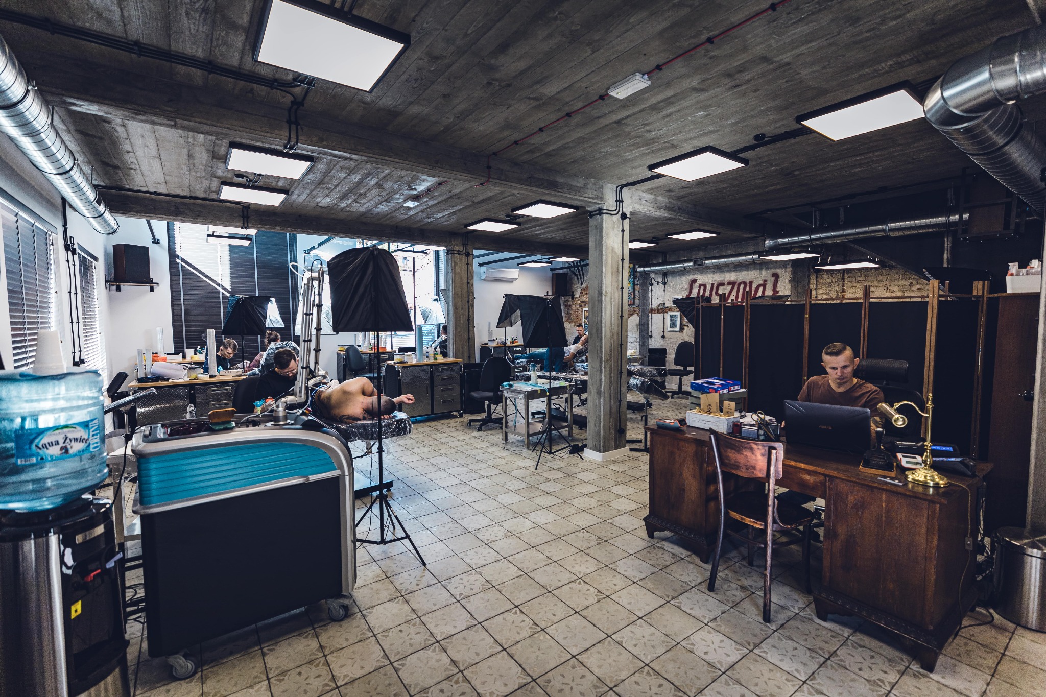 Wnętrze studia tatuażu z betonowym sufitem, widoczne stanowiska pracy z leżącymi klientami, lampami i tatuażystami w trakcie pracy, w tle biurko z pracownikiem przy komputerze.
