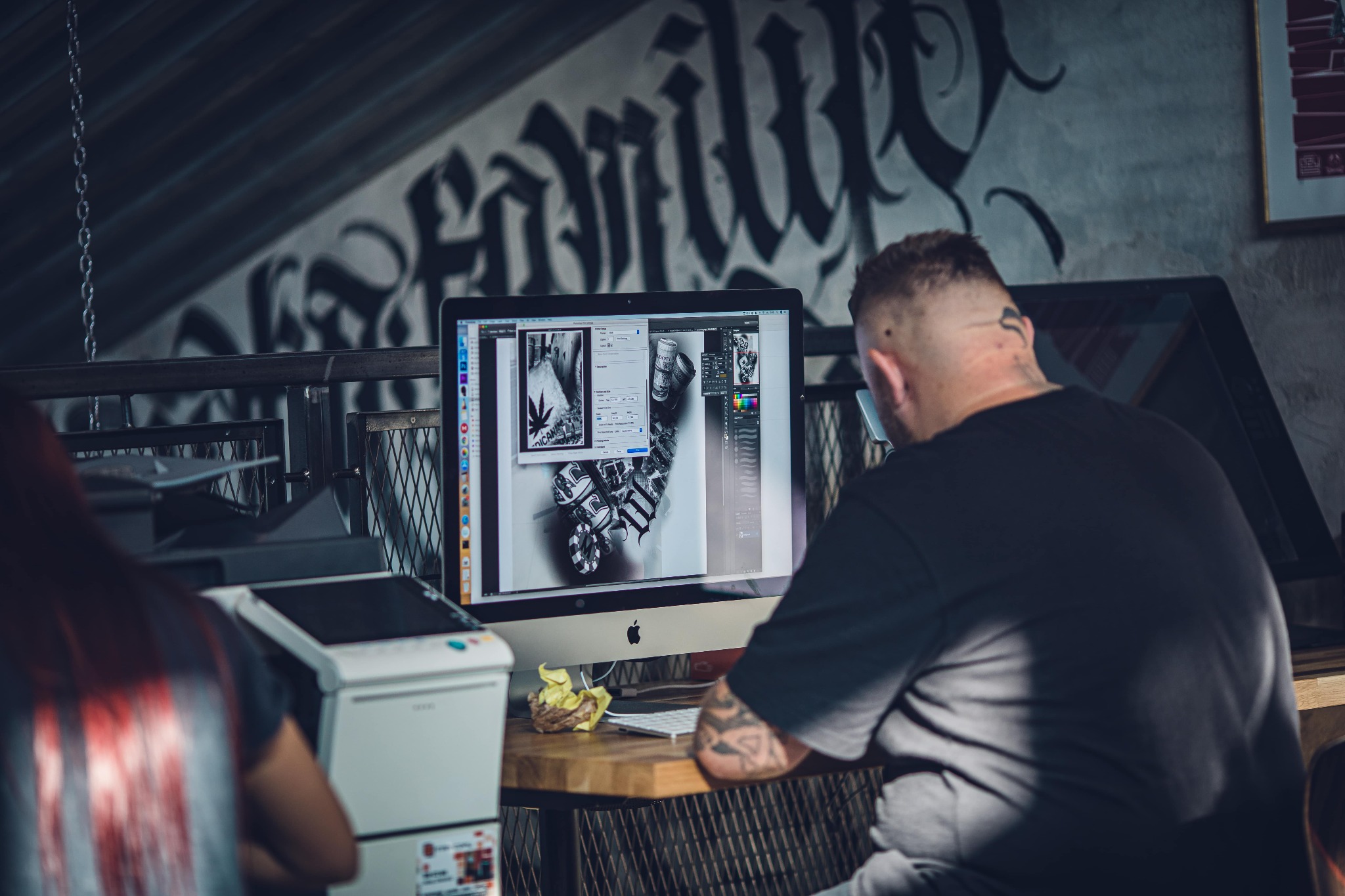 Grafik pracuje nad projektem na komputerze iMac, widoczne oprogramowanie graficzne z edytowaną grafiką w stylu tatuażu, w tle graffiti na ścianie i drukarka.