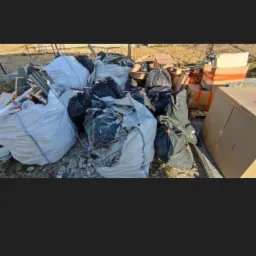 Sterta worków i kartonów z odpadami budowlanymi i gruzem na zewnątrz, widoczne elementy konstrukcji metalowej w tle.