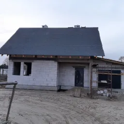 Surowy stan budynku w Toruniu z widocznym dachem pokrytym papą, mury z białego bloczka, okna i drzwi zabite czarną folią oraz rusztowanie z boku.